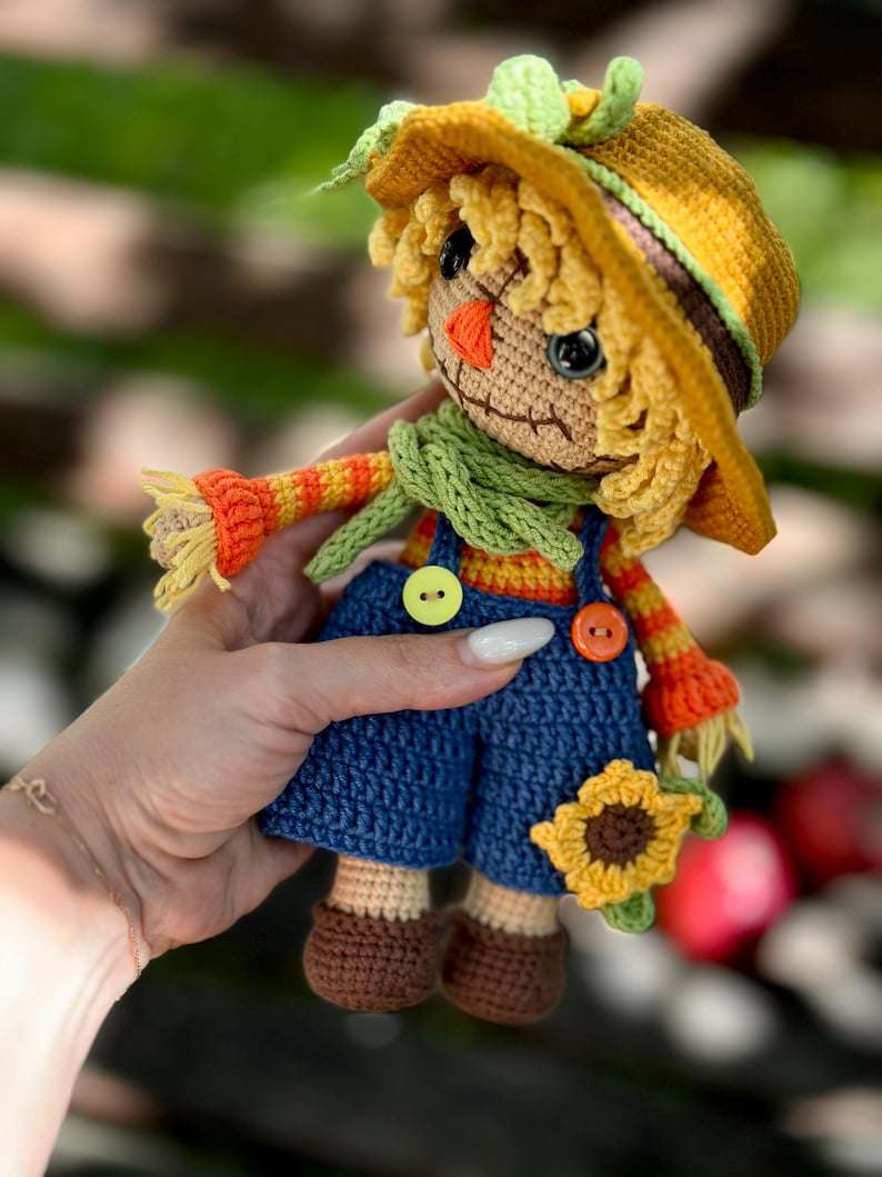 May include: A handmade crochet scarecrow doll with a yellow hat, straw hair, and a green scarf. The doll wears blue dungarees with orange and yellow striped sleeves. It holds a small sunflower. The doll is approximately 15 cm tall.