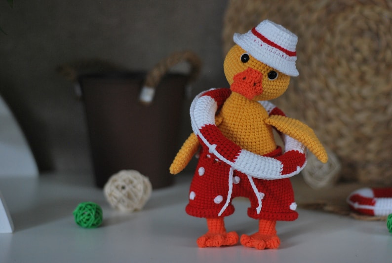 May include: A crocheted yellow duck wearing a red and white life preserver, red and white polka dot swim trunks, and a white and red striped hat.