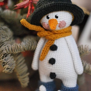 May include: A crocheted snowman toy with a black hat, a yellow scarf, and black buttons. The snowman is wearing blue boots with white accents.