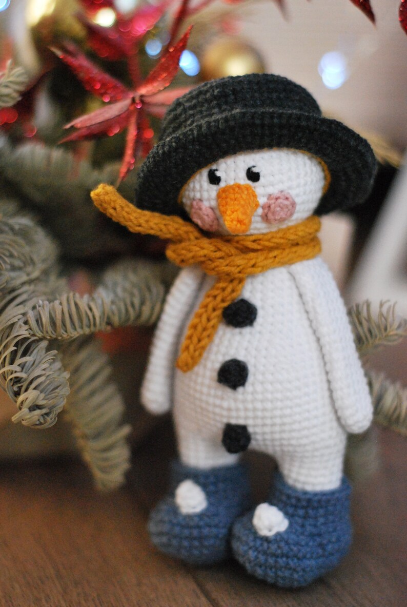May include: A crocheted snowman toy with a black hat, a yellow scarf, and black buttons. The snowman is wearing blue boots with white accents.