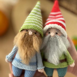 May include: Two crocheted gnomes, one with a green and white striped hat and a brown beard, the other with a red and white striped hat and a gray beard. Both gnomes are wearing blue and green outfits.