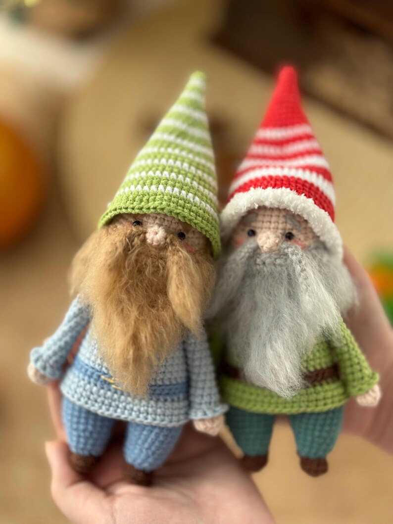 May include: Two crocheted gnomes, one with a green and white striped hat and a brown beard, the other with a red and white striped hat and a gray beard. Both gnomes are wearing blue and green outfits.