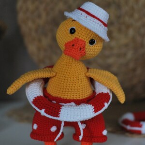 May include: A crocheted yellow duck wearing a white hat with a red stripe, red and white polka dot shorts, and a red and white life preserver.