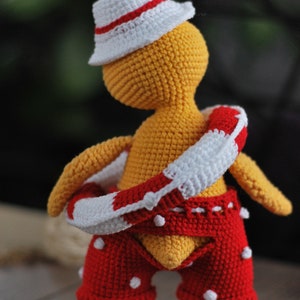 May include: A crocheted yellow toy wearing a white hat with a red stripe, red and white striped life preserver, and red shorts with white polka dots.