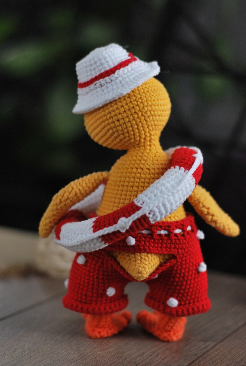 May include: A crocheted yellow toy wearing a white hat with a red stripe, red and white striped life preserver, and red shorts with white polka dots.