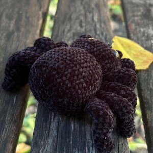 Peut inclure: Un jouet d'araign&eacute;e fait &agrave; la main, en crochet marron fonc&eacute;, avec huit pattes et un corps rond. L'araign&eacute;e en peluche est pos&eacute;e sur un banc en bois. Une feuille jaune est &agrave; proximit&eacute;.