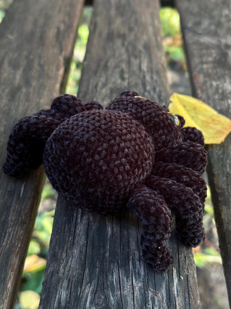 Peut inclure: Un jouet d'araign&eacute;e fait &agrave; la main, en crochet marron fonc&eacute;, avec huit pattes et un corps rond. L'araign&eacute;e en peluche est pos&eacute;e sur un banc en bois. Une feuille jaune est &agrave; proximit&eacute;.