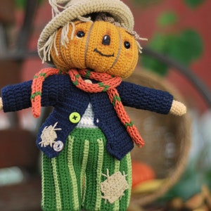 May include: A crocheted pumpkin scarecrow doll wearing a blue jacket, orange scarf, green striped trousers, and brown shoes.