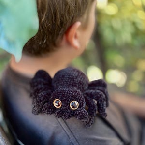 Peut inclure: Une araign&eacute;e noire au crochet avec de grands yeux ronds et huit pattes. L'araign&eacute;e est faite d'un fil doux et textur&eacute; et a des bordures d'yeux dor&eacute;es. L'araign&eacute;e repose sur une surface de couleur fonc&eacute;e.