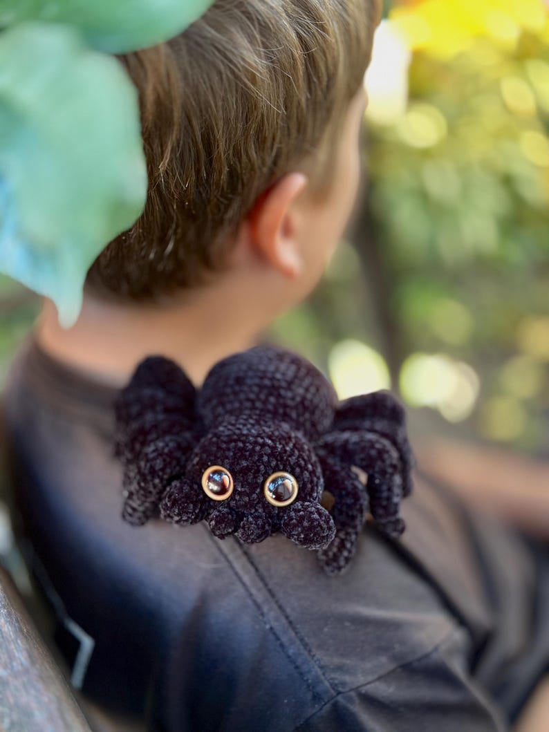 Peut inclure: Une araign&eacute;e noire au crochet avec de grands yeux ronds et huit pattes. L'araign&eacute;e est faite d'un fil doux et textur&eacute; et a des bordures d'yeux dor&eacute;es. L'araign&eacute;e repose sur une surface de couleur fonc&eacute;e.