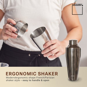 May include: A stainless steel cocktail shaker with a modern, ergonomic design. The shaker is shown in two parts, with the top and bottom sections separated. The text "ERGONOMIC SHAKER" and "Modern/ergonomic shape French/Parisian shaker style - easy to handle & open" is displayed below the shaker.