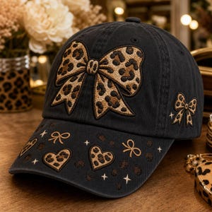 May include: A black baseball cap featuring a large embroidered leopard print bow on the front panel. The brim is decorated with smaller embroidered leopard print hearts, bows, and star accents. The side of the cap also displays a small leopard print bow.