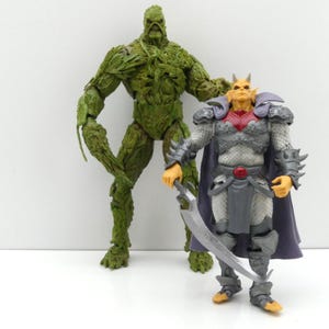 Könnte beinhalten: Zwei Actionfiguren stehen zusammen. Eine ist eine große, grüne, pflanzenartige Kreatur mit einem strukturierten, blättrigen Körper. Die andere ist eine gelbhäutige humanoide Figur in grauer Rüstung mit violettem Umhang und einem geschwungenen Schwert.