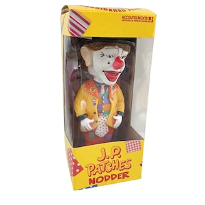 May include: A J.P. Patches bobblehead figure inside a yellow cardboard box. The clown figure has a white face, red nose, brown hair, and a yellow jacket over a colorful vest and white polka-dot tie. The box features the text J.P. PATCHES NODDER.