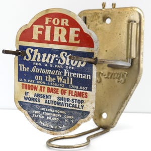 Könnte beinhalten: Eine vintage Shur-Stop automatische Feuerlöscher-Wandhalterung aus messingfarbenem Metall. Die Vorderseite zeigt ein rot-weiß-blaues Schild mit dem Text: FOR FIRE, Shur-Stop, The Automatic Fireman on the Wall, THROW AT BASE OF FLAMES.