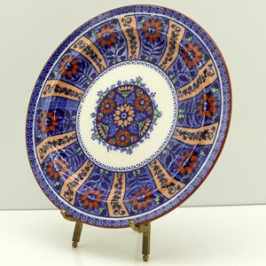 Puede incluir: Un plato de cerámica redondo que presenta un medallón circular central con motivos florales en naranja y azul. El borde consta de paneles alternos de motivos florales azules y bandas de color melocotón, enmarcado por un fino borde rojo oscuro.