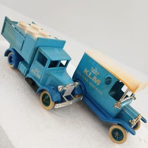May include: Two vintage blue KLM toy trucks. One truck has a flatbed with white containers, the other has a covered cargo area. Both trucks have the KLM logo and text. The trucks have cream-colored wheels and silver detailing.