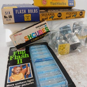 May include: A collection of vintage flashbulbs and accessories. Boxes of flashbulbs in blue, yellow, and white are visible, along with a blue plastic flash unit. The boxes have text such as "FLASH BULBS" and "SYLVANIA BLUE DOT".