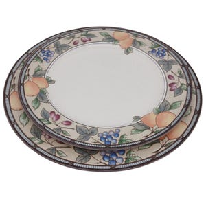 May include: Two round ceramic plates stacked on top of each other. The plates have a cream-colored center and a beige border with a fruit and leaf design. The fruit includes peaches and grapes, with a blue and brown border.