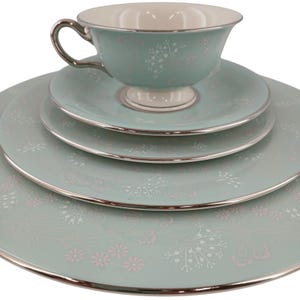 Può includere: Servizio da tè in quattro pezzi di colore verde acqua chiaro con finiture argentate. Il set comprende una tazza, un piattino e due piatti, tutti decorati con un delicato motivo floreale bianco e rosa. La tazza ha un manico argentato.