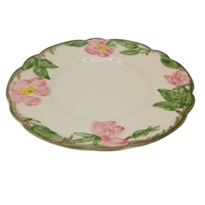 May include: A white ceramic plate with a scalloped edge, adorned with pink rose blossoms and green leaves. The plate's rim is outlined in a muted green. A classic floral design for serving food.