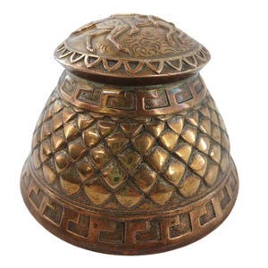 May include: A bronze-colored inkwell with a detailed, textured design. The inkwell features a lid with a decorative border and a relief of figures. The body has a diamond pattern and a Greek key border, showcasing an antique aesthetic.