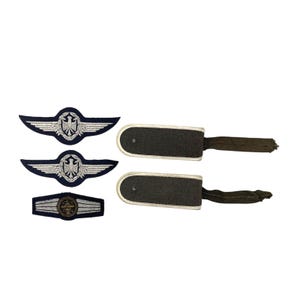 May include: A selection of vintage military insignia. The image shows three embroidered wing badges with eagle emblems and two dark grey shoulder boards with olive green straps. The badges have silver and blue detailing.
