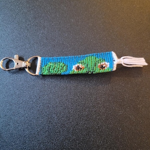 May include: A blue and green woven keyring with a white tassel. The keyring features a design of two green frogs with white eyes.