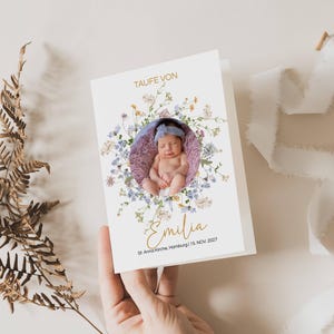 Pode incluir: Um cartão branco com um design de coroa de flores e uma foto de uma menina pequena no centro. O texto "Taufe von Emilia" está escrito em letras douradas abaixo da foto. A data "18. NOV. 2022" está escrita em letras pretas abaixo do nome.