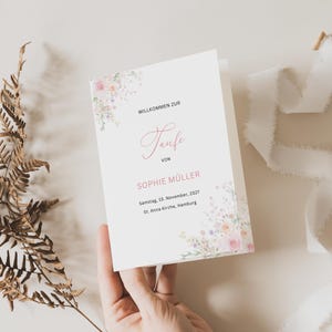 May include: A white folded invitation with floral watercolor accents. The text reads "WILLKOMMEN ZUR Taufe VON SOPHIE MÜLLER Samstag, 15. November, 2027 St. Anna Kirche, Hamburg."