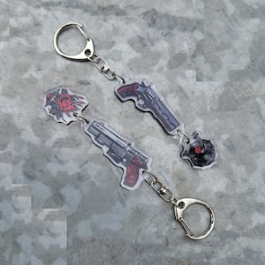 Puede incluir: Dos llaveros de acrílico transparente con cadenas plateadas. Un llavero presenta una pistola negra y plateada con mango rojo y un diseño de llama roja y negra. El otro llavero presenta un revólver negro y plateado con mango rojo y un diseño de calavera negra y roja.