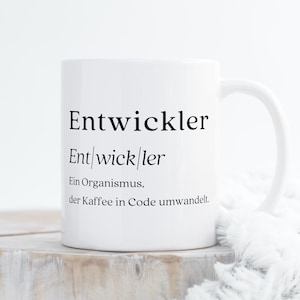 May include: White ceramic mug with black text that reads "Entwickler" and a definition in German. The definition reads "Ent/wick/ler Ein Organismus, der Kaffee in Code umwandelt."