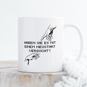 以下が含まれることがあります： 白いセラミック製のマグカップに、黒色の2つの手のグラフィックが描かれています。片方は指を指し示し、もう片方は開いています。マグカップには「HABEN SIE ES MIT EINEM NEUSTART VERSUCHT?」というテキストが書かれています。
