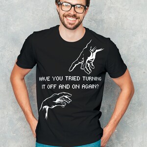 Może przedstawiać: Czarny t-shirt z białym nadrukiem przedstawiającym dwie ręce sięgające do siebie. Pod nadrukiem znajduje się tekst "Have you tried turning it off and on again?"