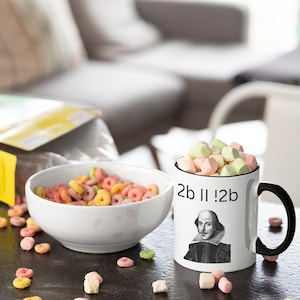 Op de afbeelding: Een witte mok met een zwart handvat en een zwarte rand. De mok heeft een zwart-witte afbeelding van William Shakespeare met de tekst "2b || !2b" erop gedrukt. De mok is gevuld met pastelkleurige marshmallows en staat op een zwart oppervlak met verspreide ontbijtringen en marshmallows.