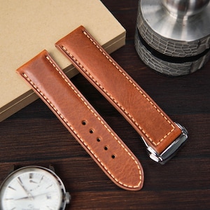 May include: Two brown leather watch straps with cream stitching. One strap has five holes for the buckle. The straps are on a dark wooden surface. A silver watch face is visible in the lower left corner.