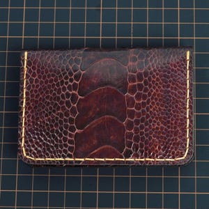 May include: A brown leather card holder with a textured ostrich leg pattern. The edges are stitched with gold thread. The card holder is rectangular and has a dark brown color.