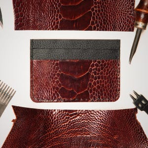 Peut inclure: Un porte-cartes rectangulaire composé d'une section inférieure en cuir rouge foncé à motif autruche texturé et d'une bande supérieure en cuir noir lisse, entouré de chutes de cuir et d'outils de maroquinerie sur fond blanc.