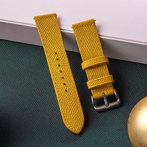 May include: A mustard yellow watch strap with a textured grain finish, featuring two matching leather keepers and a polished silver-tone metal buckle. The strap has a series of punched holes for adjustment and visible tonal stitching along the edges.