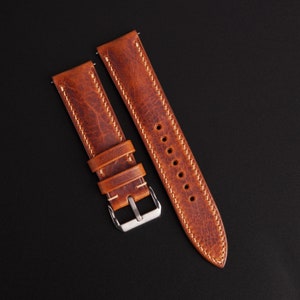 Pode incluir: Uma pulseira de relógio de duas peças feita de couro castanho desgastado com uma superfície texturizada. A pulseira apresenta costuras de cor clara ao longo das bordas, dois passadores de couro e uma fivela de metal prateado polido.