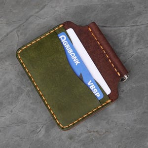 Puede incluir: Cartera de cuero hecha a mano en verde oliva y marrón, con costuras visibles. Contiene una tarjeta Agribank azul y una tarjeta blanca. Diseño minimalista con cierre seguro.