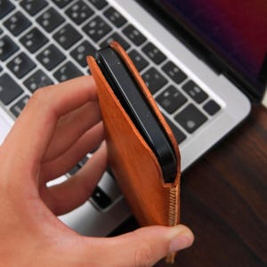 May include: A slim, tan leather sleeve case holding a black smartphone. The sleeve features visible light-colored stitching along the side edge, providing a secure fit for the device while maintaining a simple, functional design for mobile protection.