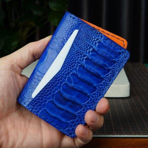 Pode incluir: Um porta-cartões azul brilhante com um padrão de couro de pata de avestruz texturizado com protuberâncias e escamas visíveis. A carteira tem uma ranhura vertical com um cartão branco e um forro interior laranja na borda superior.
