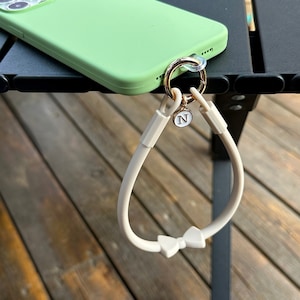 May include: A white phone strap with a bow detail and a gold charm with the letter 'N' on it. The strap is attached to a green phone case.