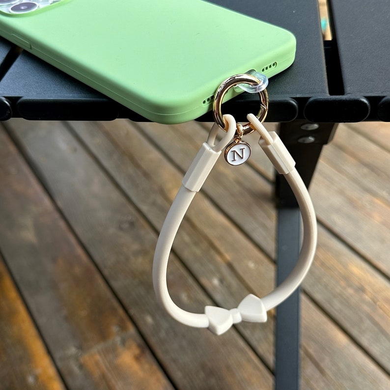 May include: A white phone strap with a bow detail and a gold charm with the letter 'N' on it. The strap is attached to a green phone case.