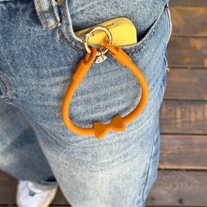 May include: A bright orange silicone phone strap with a bow detail. The strap is attached to a gold key ring and is hanging from a pair of blue jeans.