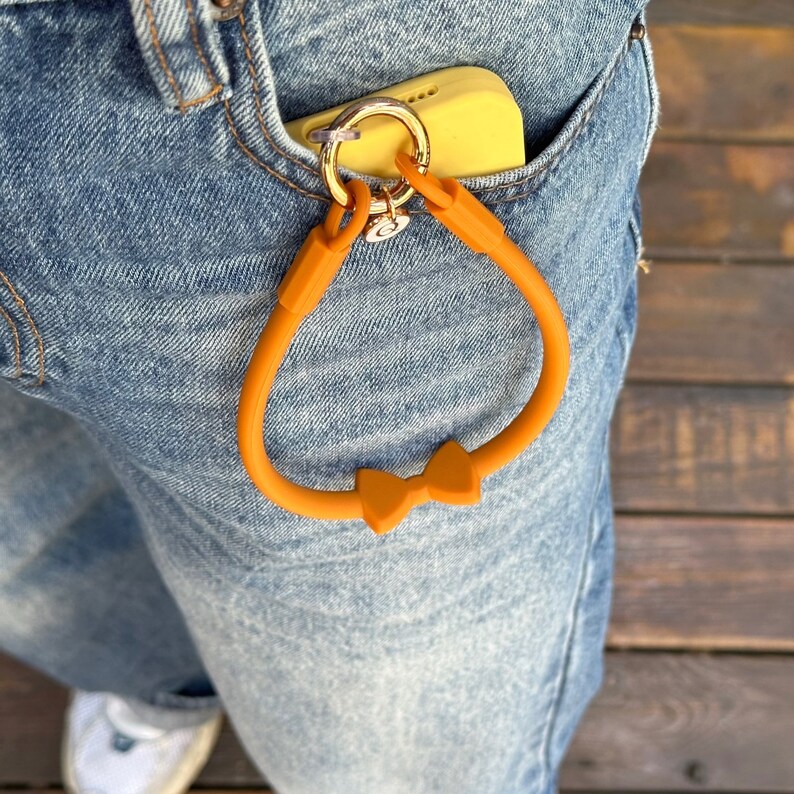 May include: A bright orange silicone phone strap with a bow detail. The strap is attached to a gold key ring and is hanging from a pair of blue jeans.