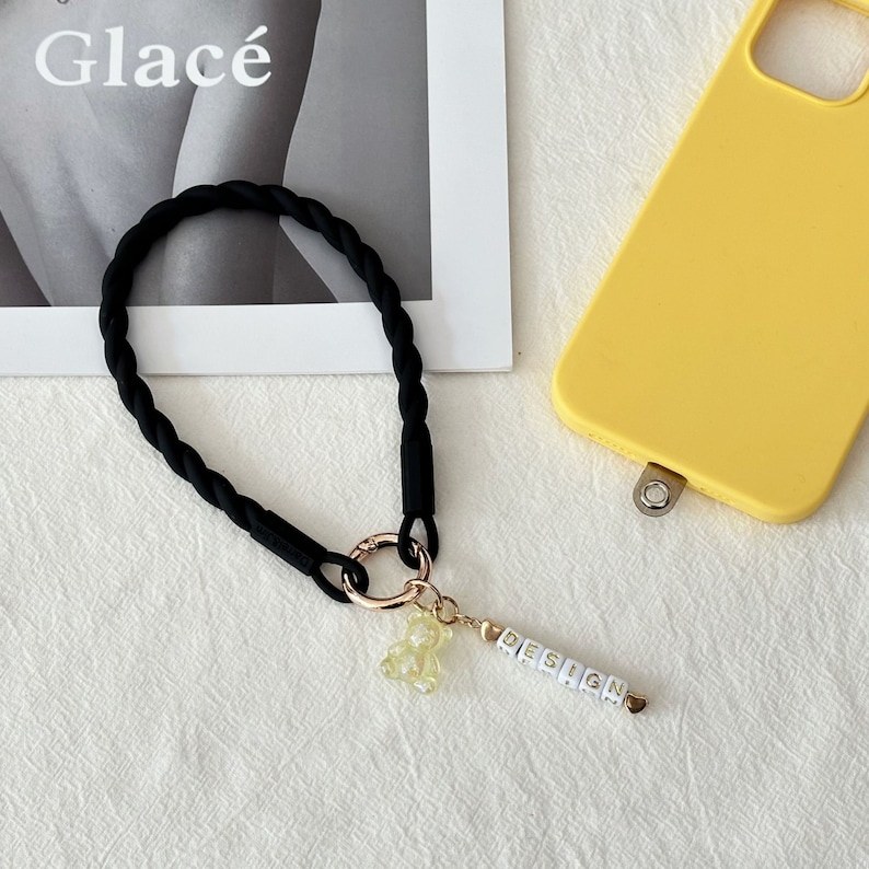 May include: Black braided phone strap with a gold ring and a charm featuring a yellow gummy bear and the word "DESIGN" spelled out in white beads.