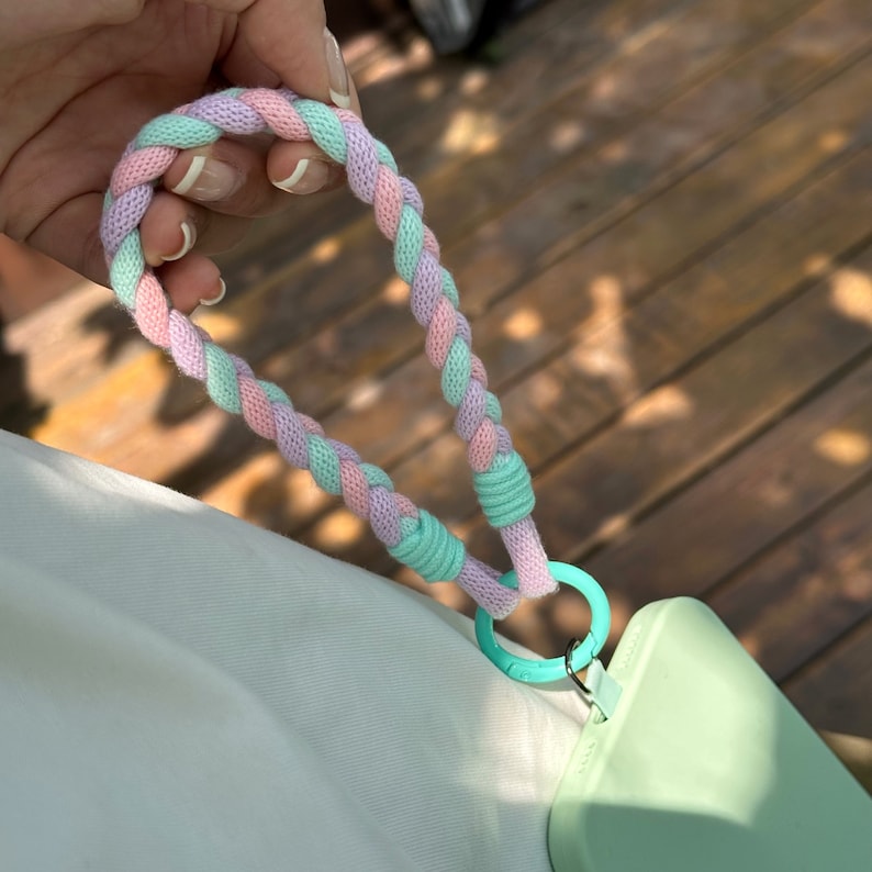 K&ouml;nnte beinhalten: Eine hellgr&uuml;ne Handyh&uuml;lle mit einem rosa und hellgr&uuml;nen geflochtenen Band und einem silbernen Ring.