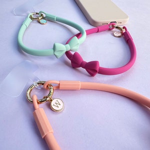 May include: Three silicone phone straps with bow accents. The straps are in mint green, pink, and peach. Each strap has a gold charm with a letter on it. The charms are "W", "A", and a small circle.